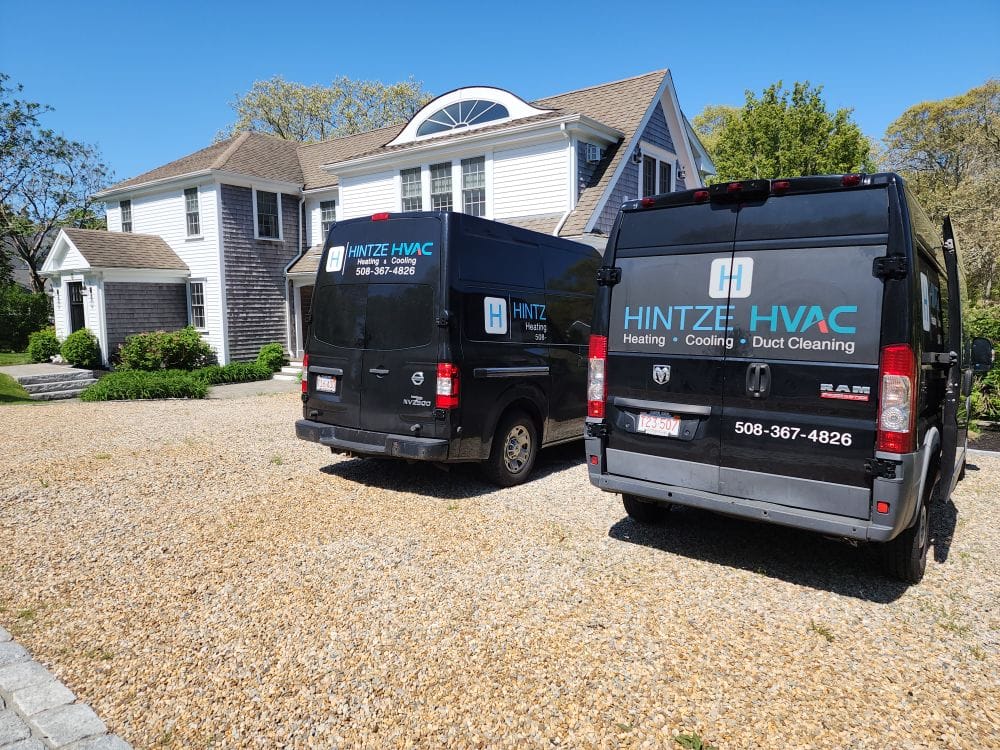497547163_2091412317994742_3247179955237981458_n Two Hintze HVAC vans parked on a gravel driveway in front of a white house on a sunny day.